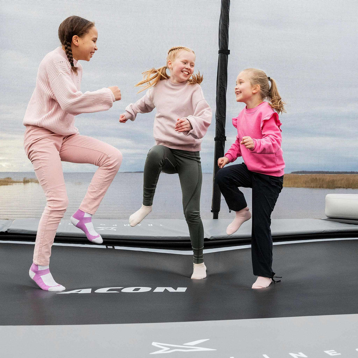 Young children in vibrant pink clothing enjoying active play time on their Acon X Trampoline, demonstrating safe recreational usage and family-friendly design.