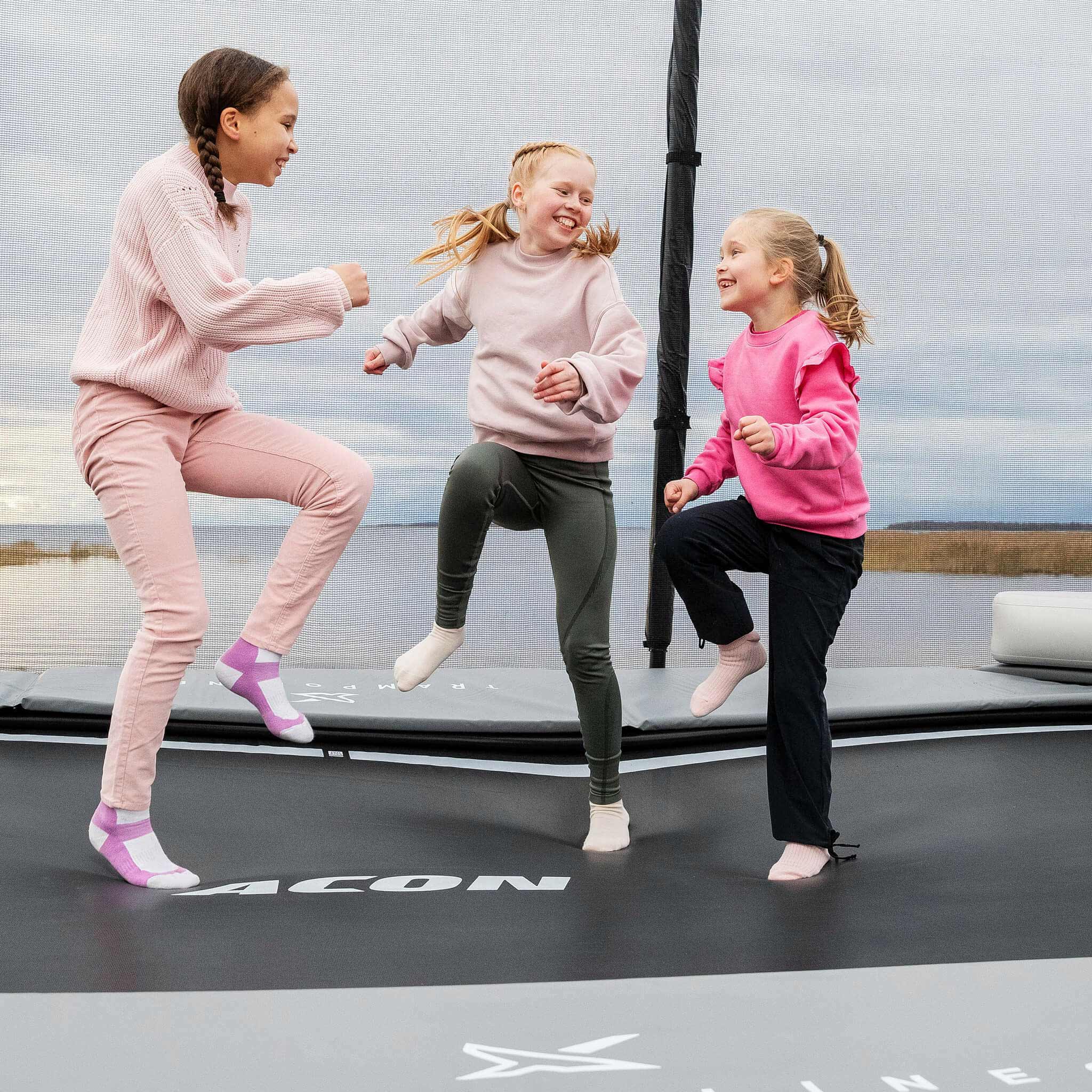 Young children in vibrant pink clothing enjoying active play time on their Acon X Trampoline, demonstrating safe recreational usage and family-friendly design.