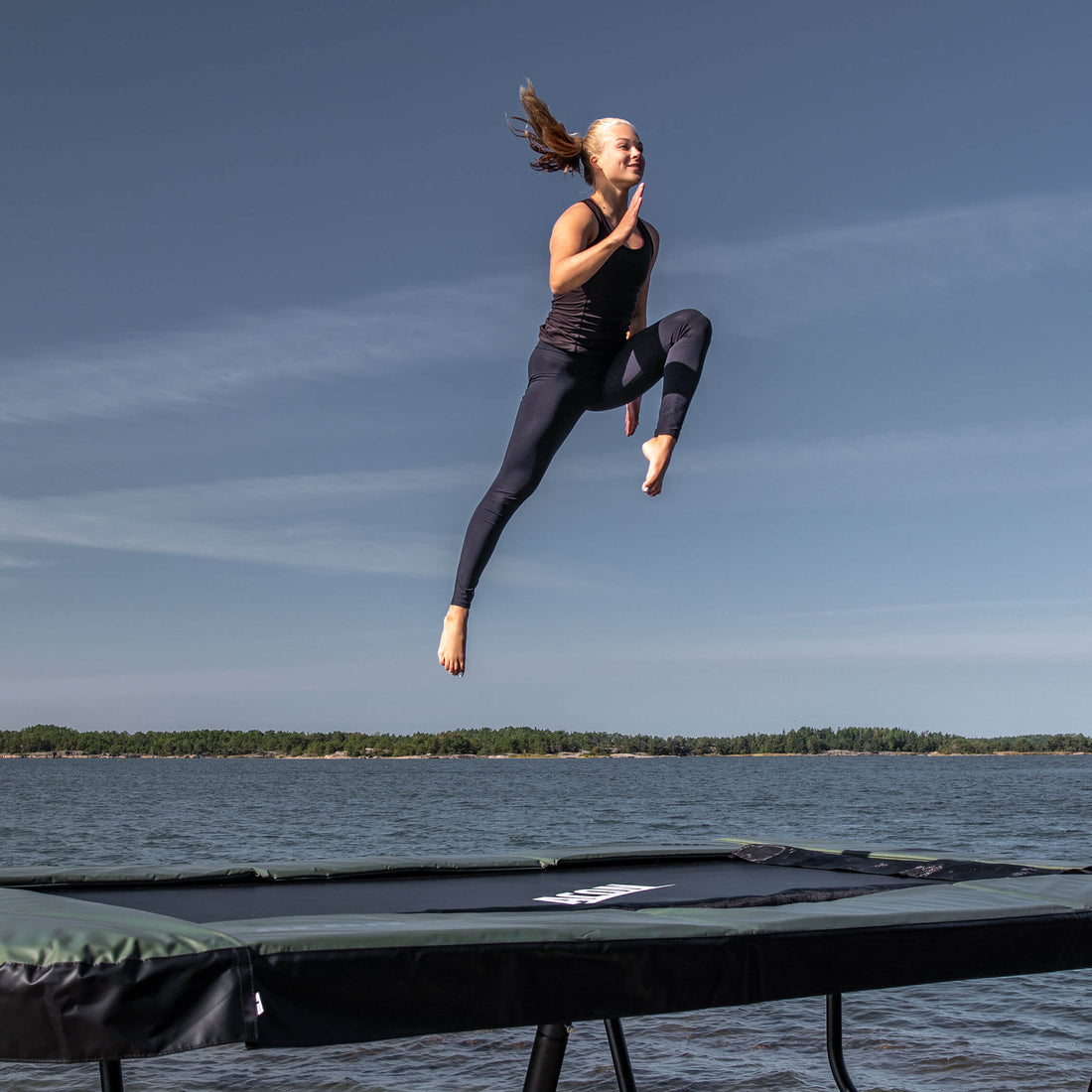 Professional athlete demonstrating advanced aerial techniques on an Acon 13 HD trampoline against scenic coastal landscape, highlighting performance capabilities for serious training applications.