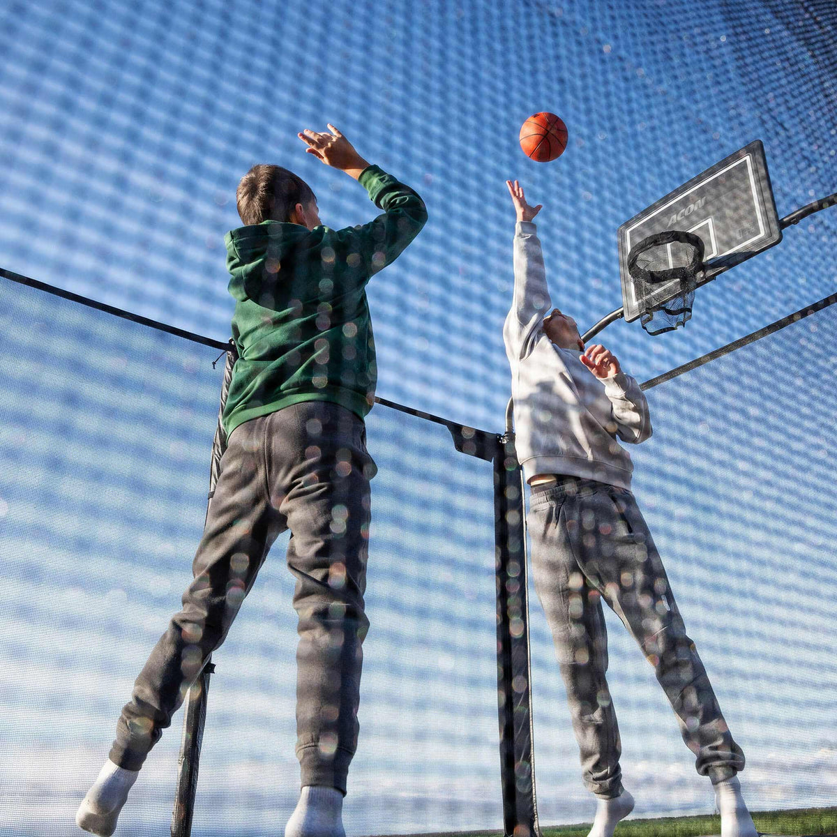 Recreational basketball activity on the Acon X Trampoline featuring young athletes combining bouncing with sport skills, demonstrating versatile entertainment possibilities.