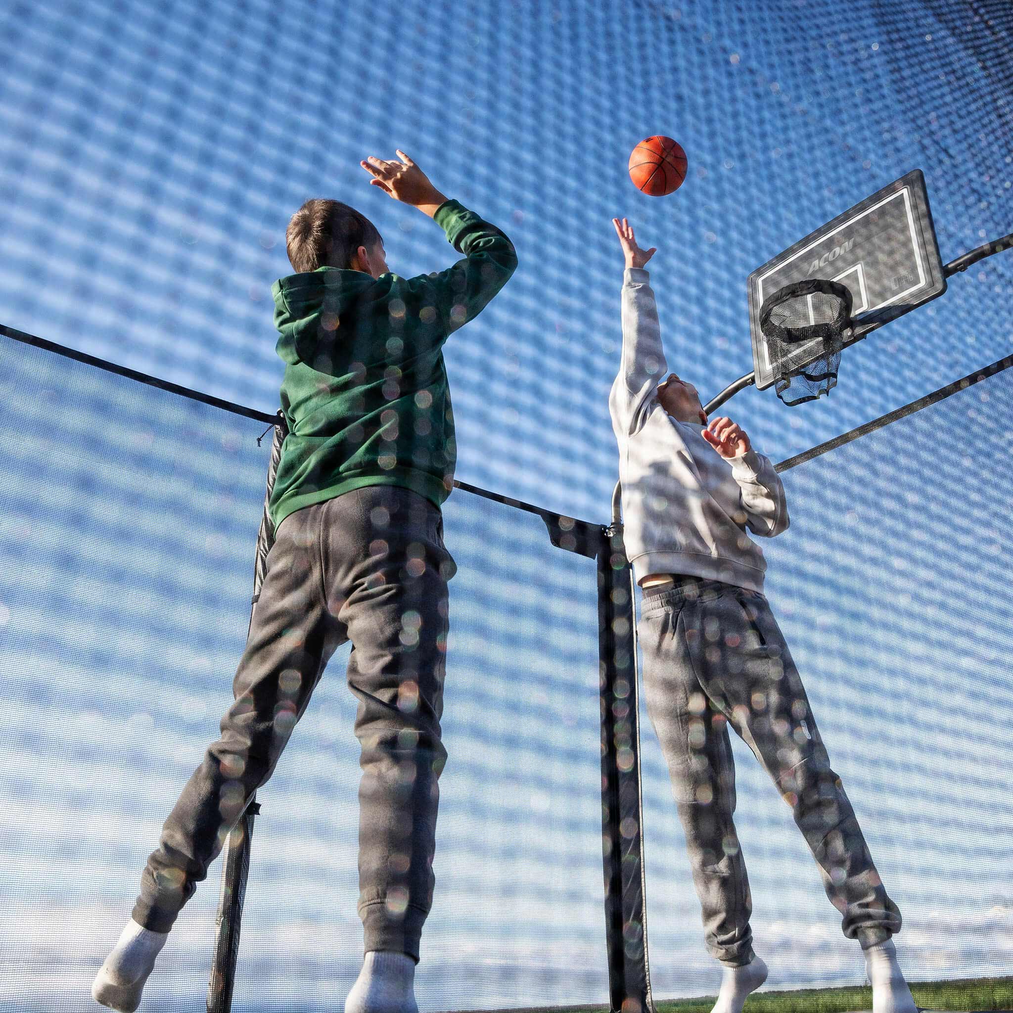 Recreational basketball activity on the Acon X Trampoline featuring young athletes combining bouncing with sport skills, demonstrating versatile entertainment possibilities.