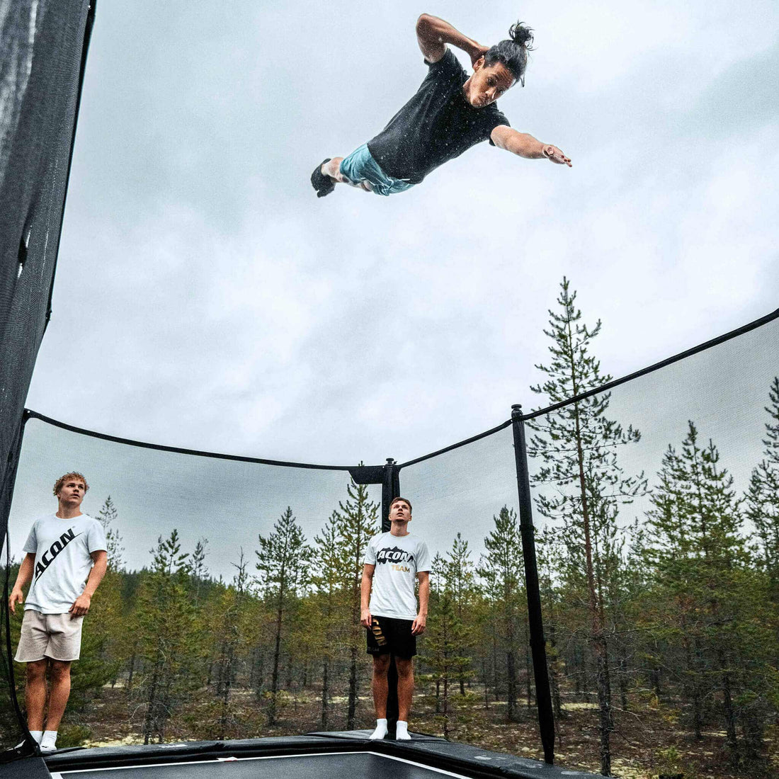Skilled performer demonstrating advanced techniques on an Acon X Trampoline while companions watch, showcasing the equipment's capability for serious athletic training and entertainment.