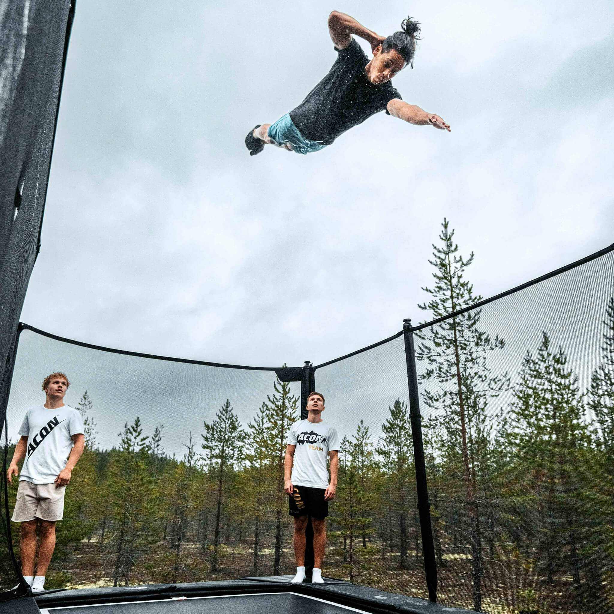 Skilled performer demonstrating advanced techniques on an Acon X Trampoline while companions watch, showcasing the equipment's capability for serious athletic training and entertainment.